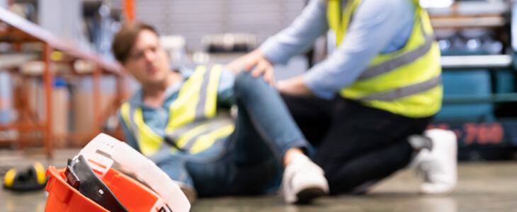 Worker assisting injured colleague on the floor, hard hat in the foreground.