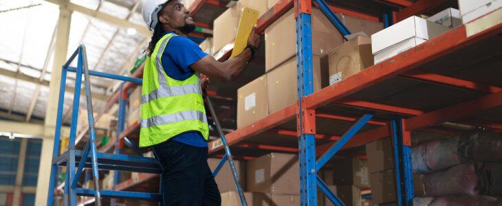 Warehouse worker standing on ladder checking inventory on shelf.